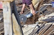 A person is using a power tool to cut through metal rods on a construction site. Sparks are flying from the point of contact between the tool and the metal, and the area is filled with various construction materials like rebar and wooden planks. The person is wearing tan pants and a purple shirt.