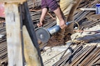 A person is using a power tool to cut through metal rods on a construction site. Sparks are flying from the point of contact between the tool and the metal, and the area is filled with various construction materials like rebar and wooden planks. The person is wearing tan pants and a purple shirt.