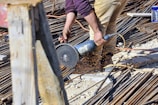 A person is using a power tool to cut through metal rods on a construction site. Sparks are flying from the point of contact between the tool and the metal, and the area is filled with various construction materials like rebar and wooden planks. The person is wearing tan pants and a purple shirt.