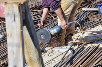 A person is using a power tool to cut through metal rods on a construction site. Sparks are flying from the point of contact between the tool and the metal, and the area is filled with various construction materials like rebar and wooden planks. The person is wearing tan pants and a purple shirt.