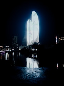 A futuristic skyline features a set of illuminated, glass skyscrapers with curved, organic forms. The buildings emit a soft blue and white light, reflecting beautifully onto the surface of a nearby river. Surrounding structures, though less prominent, suggest an urban setting. The scene exudes a sense of modernity and technological advancement.