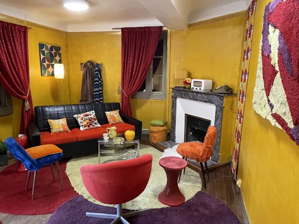 A cozy living room with vibrant, eclectic decor. The mustard yellow walls are complemented by red curtains. There's a black leather sofa adorned with colorful cushions, and a glass coffee table in front. A mix of chairs in blue and red surround the table. A fireplace is set against the wall, topped with a retro radio and a flower vase. Various textured rugs cover the wooden floor, and there's wall art and a modern lamp adding to the room's character.
