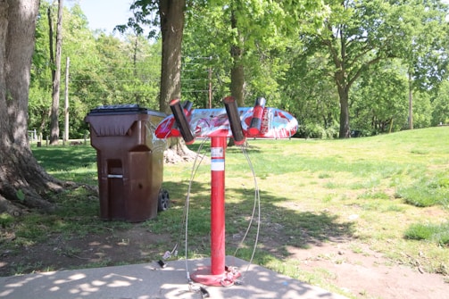 A vibrant red bike repair station stands in a park with lush green grass and trees. To the left of the station is a large brown trash bin. Sunlight casts shadows on the concrete pathway, creating a serene outdoor setting.