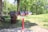 A vibrant red bike repair station stands in a park with lush green grass and trees. To the left of the station is a large brown trash bin. Sunlight casts shadows on the concrete pathway, creating a serene outdoor setting.