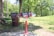 A vibrant red bike repair station stands in a park with lush green grass and trees. To the left of the station is a large brown trash bin. Sunlight casts shadows on the concrete pathway, creating a serene outdoor setting.