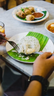 A close-up view of a plate featuring a serving of white rice garnished with herbs, placed on a banana leaf. A hand holding a fork is seen scooping the rice. In the background, there is another dish with spring rolls and a bowl of soup.