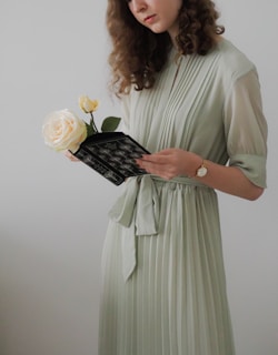 A woman in a light green pleated dress holds a small bouquet consisting of a cream-colored rose and a smaller yellow rose while reading a black book with decorative designs. She has curly brown hair and wears a watch on her left wrist.
