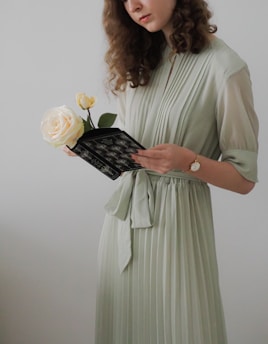 A woman in a light green pleated dress holds a small bouquet consisting of a cream-colored rose and a smaller yellow rose while reading a black book with decorative designs. She has curly brown hair and wears a watch on her left wrist.