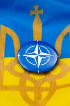 A blue circular badge with a white compass rose symbol is centered on a flag with horizontal stripes of blue and yellow, incorporating an abstract trident design.