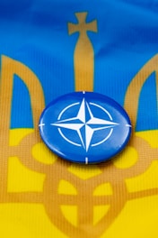 A blue circular badge with a white compass rose symbol is centered on a flag with horizontal stripes of blue and yellow, incorporating an abstract trident design.