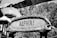 A rustic, woven sign with detailed carvings and the word 'ALOHA!' stands prominently. Surrounding it are tropical elements, including thatched roof structures with intricate patterns and lush vegetation in the background.