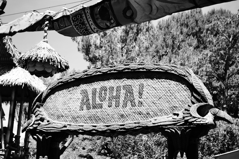 A rustic, woven sign with detailed carvings and the word 'ALOHA!' stands prominently. Surrounding it are tropical elements, including thatched roof structures with intricate patterns and lush vegetation in the background.