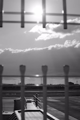 A black and white photograph depicting an airport tarmac. In the foreground, a fence is in focus with vertical lines and a blurred view of a jet bridge leading to the tarmac. The background shows a row of industrial buildings set against a dramatic sky with sun rays breaking through the clouds.