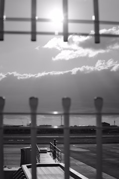 A black and white photograph depicting an airport tarmac. In the foreground, a fence is in focus with vertical lines and a blurred view of a jet bridge leading to the tarmac. The background shows a row of industrial buildings set against a dramatic sky with sun rays breaking through the clouds.