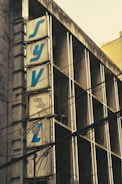 A building facade with large vertical signage displaying blue letters against a weathered, beige background. The structure shows signs of aging with visible dark streaks on the walls. Electrical wires cross the view, adding a sense of urban clutter and complexity. The architectural style appears utilitarian with a focus on function over ornamentation.