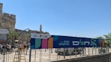 A large outdoor banner with colorful abstract shapes is displayed in front of an ancient stone wall. Several people are gathered around, and a crane is positioned nearby. The sky is clear and blue, indicating a sunny day.