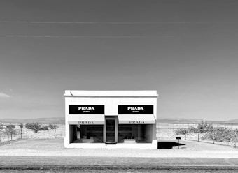 A standalone building resembling a storefront in a desert landscape with clear skies. The facade displays the Prada logo above two large windows that showcase merchandise. Sparse vegetation is visible in the background, and the minimalistic setting emphasizes isolation and simplicity.