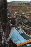 A person is climbing a large rock formation in an outdoor setting, using both hands for grip. They are wearing a green sweater and appear focused on the climb. Below, there is a pad labeled 'SNAP' for protection, set amidst a rugged landscape with scattered rocks and a distant view of rolling hills and a road with a few cars.