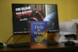 A book titled 'Do'a & Wirid' is prominently placed on a desk in front of a computer monitor. The book cover features a gradient of blue to orange with yellow text, and the desk is cluttered with various objects including a keyboard and mouse. The monitor in the background displays unreadable text with a blurred image.