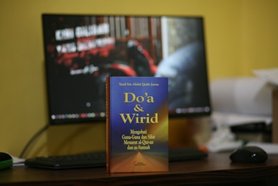 A book titled 'Do'a & Wirid' is prominently placed on a desk in front of a computer monitor. The book cover features a gradient of blue to orange with yellow text, and the desk is cluttered with various objects including a keyboard and mouse. The monitor in the background displays unreadable text with a blurred image.