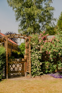 A wooden garden gate with lattice design stands open, bordered by lush climbing plants featuring pink and white flowers. Behind it, a tree towers over a charming residential area with light brown buildings. The foreground shows a neatly trimmed lawn with purple flowers at the base of the trellis.
