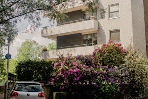 A modern apartment building with multiple balconies, surrounded by lush greenery and vibrant flowering bushes. A silver car is parked at the entrance, partially shaded by surrounding trees.