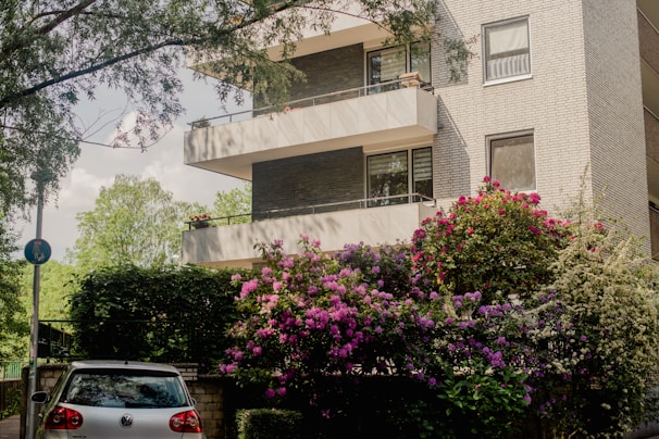 A modern apartment building with multiple balconies, surrounded by lush greenery and vibrant flowering bushes. A silver car is parked at the entrance, partially shaded by surrounding trees.
