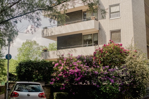 A modern apartment building with multiple balconies, surrounded by lush greenery and vibrant flowering bushes. A silver car is parked at the entrance, partially shaded by surrounding trees.