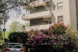 A modern apartment building with multiple balconies, surrounded by lush greenery and vibrant flowering bushes. A silver car is parked at the entrance, partially shaded by surrounding trees.