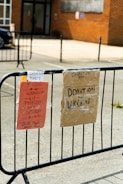 Two signs are attached to a black metal fence. The first sign on the left is orange and details opening times on Tuesday, Wednesday, Thursday, and Saturday. The second sign on the right is cardboard, labeled for donations to Ukraine. The background features a brick building and a parked car.