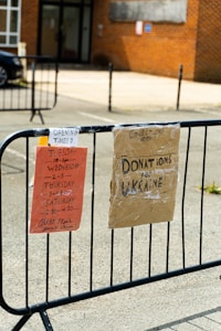 Two signs are attached to a black metal fence. The first sign on the left is orange and details opening times on Tuesday, Wednesday, Thursday, and Saturday. The second sign on the right is cardboard, labeled for donations to Ukraine. The background features a brick building and a parked car.