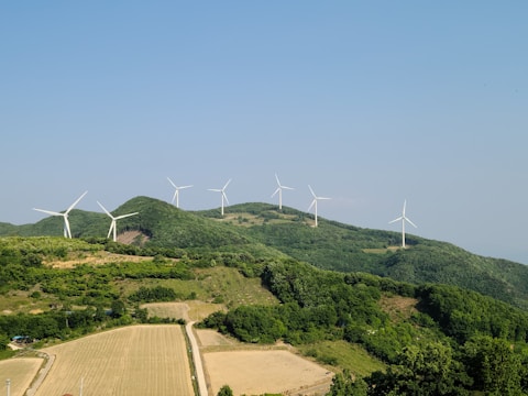 A range of wind turbines scattered across lush green hills under a clear blue sky. The turbines are positioned along the ridges of the hills, surrounded by dense forests and fields. The landscape is expansive, emphasizing the scale of the wind farm.
