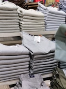 Stacks of neatly folded clothing items, primarily pants, are organized on a display shelf. The clothing items vary in different shades of gray, beige, and other muted tones. Visible tags are attached to the clothes, and a hand can be seen holding one piece of clothing.