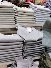 Stacks of neatly folded clothing items, primarily pants, are organized on a display shelf. The clothing items vary in different shades of gray, beige, and other muted tones. Visible tags are attached to the clothes, and a hand can be seen holding one piece of clothing.