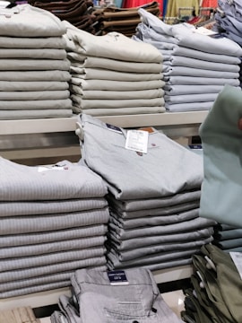 Stacks of neatly folded clothing items, primarily pants, are organized on a display shelf. The clothing items vary in different shades of gray, beige, and other muted tones. Visible tags are attached to the clothes, and a hand can be seen holding one piece of clothing.