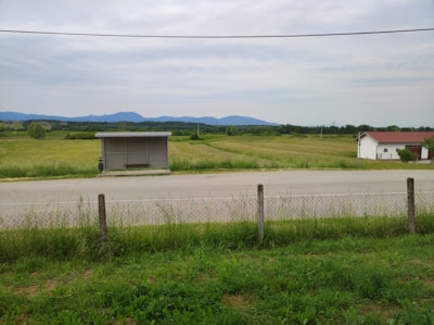 A rural landscape featuring a grassy field with a narrow road in the foreground. A metal bus stop shelter is positioned beside the road, and a small white house with a red roof is visible in the distance. Lush green fields stretch toward a series of low mountains on the horizon under an overcast sky. A fence runs parallel to the road, separating the field from the viewer.