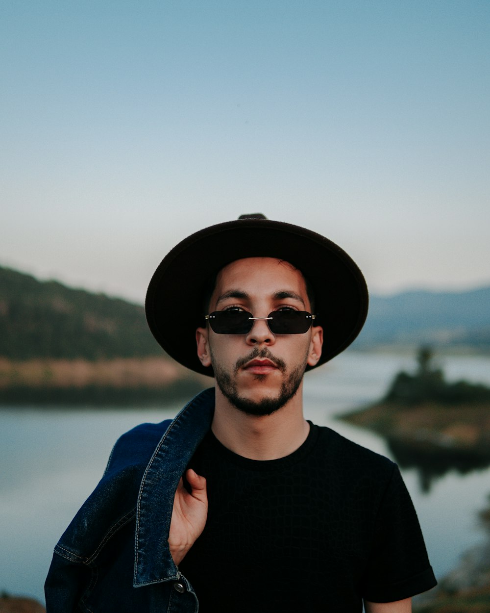 a man wearing a hat and sunglasses standing in front of a lake