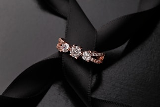 A sparkling rose gold ring with three prominent diamonds sits elegantly on a black satin ribbon background. The combination of the shiny diamonds and the luxurious texture of the ribbon creates a sophisticated and elegant presentation.