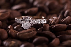 A sparkling diamond ring is set against a background of roasted coffee beans, creating an intriguing contrast between the elegance of the jewelry and the rustic texture of the beans.