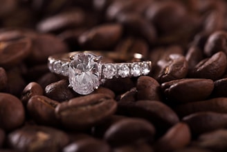 A sparkling diamond ring is set against a background of roasted coffee beans, creating an intriguing contrast between the elegance of the jewelry and the rustic texture of the beans.