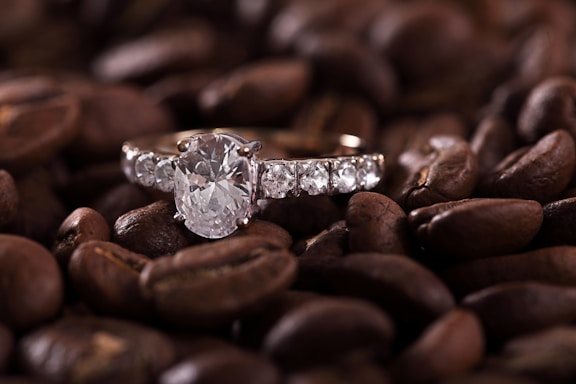 A sparkling diamond ring is set against a background of roasted coffee beans, creating an intriguing contrast between the elegance of the jewelry and the rustic texture of the beans.