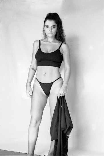 A woman stands confidently, wearing a black two-piece outfit. She holds a piece of clothing in her left hand and has a tattoo on her right arm. The image is in black and white, with a simple backdrop.