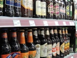 A supermarket shelf displaying various bottles of beer. The bottles feature different labels and brands, arranged in two rows. Each bottle has a price tag in front of it indicating its cost.