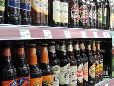 A supermarket shelf displaying various bottles of beer. The bottles feature different labels and brands, arranged in two rows. Each bottle has a price tag in front of it indicating its cost.
