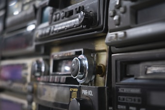 A close-up view of vintage car stereo systems, featuring various dials, buttons, and a prominent volume knob. Several of the units are stacked together, exhibiting a range of designs typical of older audio equipment.