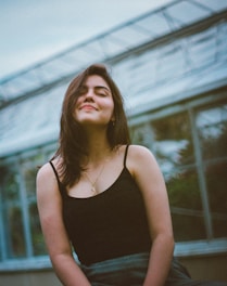 A young woman with long dark hair is smiling gently with her eyes closed. She is wearing a black sleeveless top and a necklace, sitting in front of a steel structure, possibly a greenhouse. The background is blurred with hints of greenery.