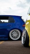 A close-up view of two modified cars showcasing custom wheels and lowered suspensions, parked on an asphalt surface. The blue car has a sleek design with a polished metal rim while a portion of a yellow car is visible in the foreground.