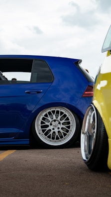 A close-up view of two modified cars showcasing custom wheels and lowered suspensions, parked on an asphalt surface. The blue car has a sleek design with a polished metal rim while a portion of a yellow car is visible in the foreground.