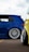 A close-up view of two modified cars showcasing custom wheels and lowered suspensions, parked on an asphalt surface. The blue car has a sleek design with a polished metal rim while a portion of a yellow car is visible in the foreground.
