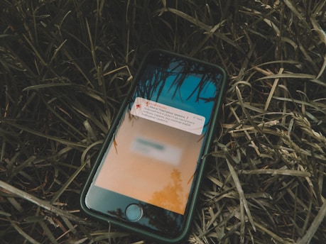 A smartphone lies on grass, displaying a notification on its screen. The background wallpaper on the phone is an abstract design with shades of blue and orange, and part of the screen is slightly blurred.